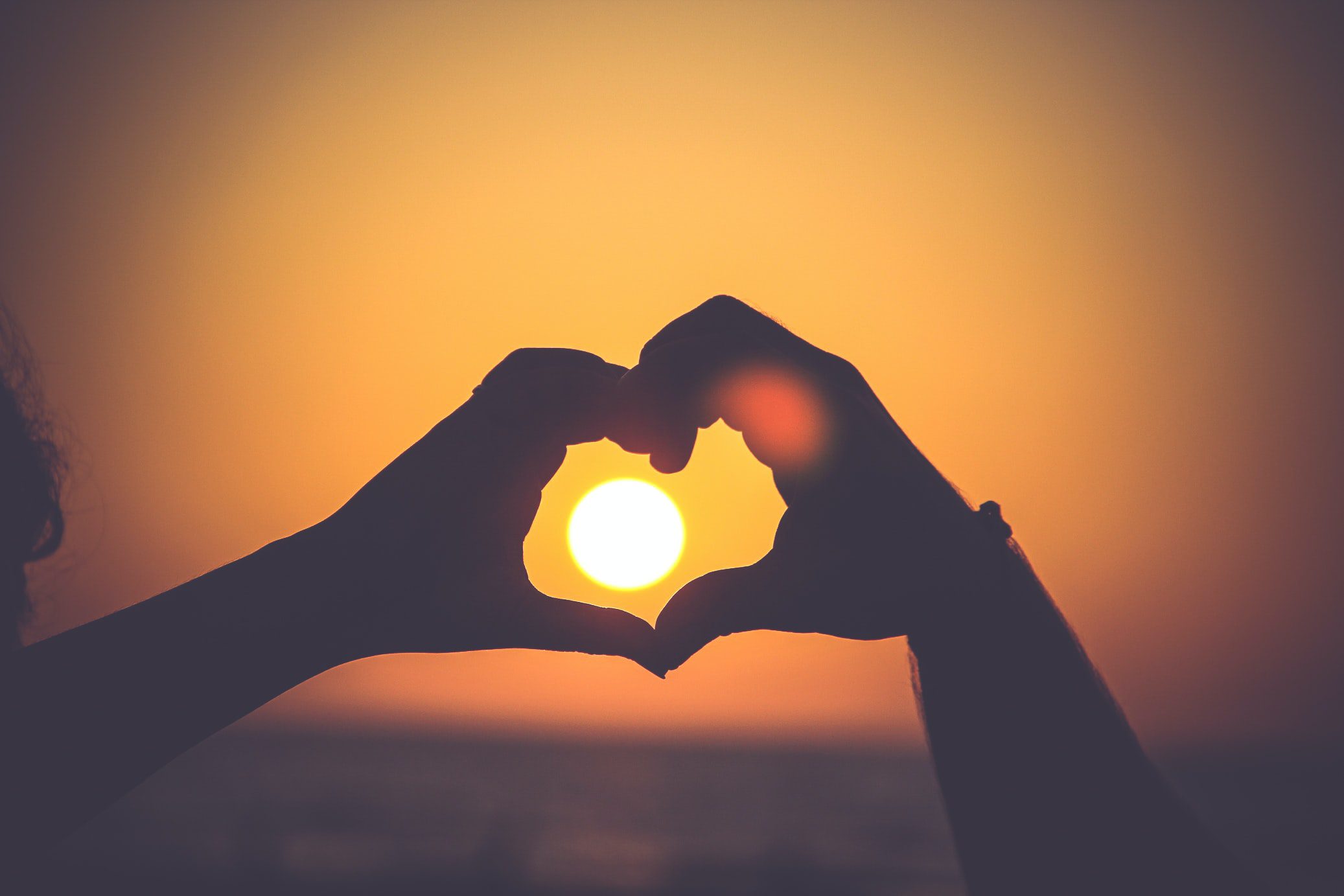 two hands forming a heart shape in front of the setting sun